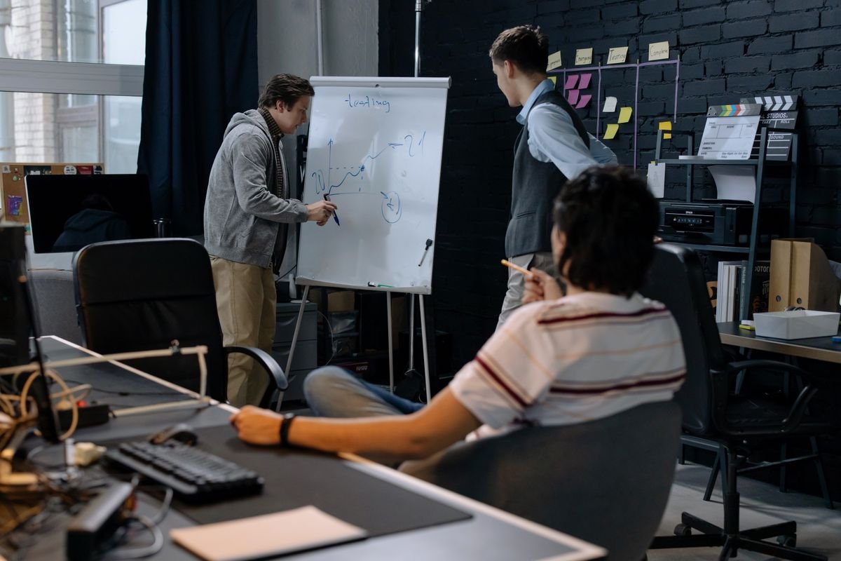 Man sketching a line graph on a flip-chart whiteboard while two colleagues watch in an office with sticky notes on a dark brick wall